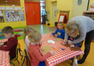 Dzieci siedzi przy stoliku, Pani Magdalena Siupa pomaga dziewczynce odrysować dłoń na kartcę papieru.